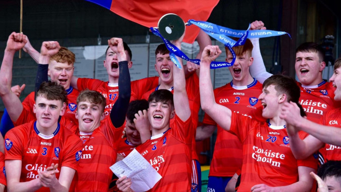 Schülerinnen und Schüler von Coláiste Choilm feiern ihren Sieg in roten Sportuniformen mit einer blauen und weißen Fahne.