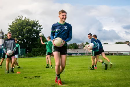 Eine Gruppe junger Athletinnen und Athleten spielt auf dem Sportfeld der Coláiste Chríost Rí vor Bäumen und Gebäuden.
