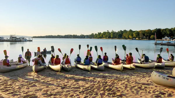 Schüler der Coláiste Éamann Rís paddeln in traditioneller Kleidung mit Kanus auf einem ruhigen See umgeben von Wald.