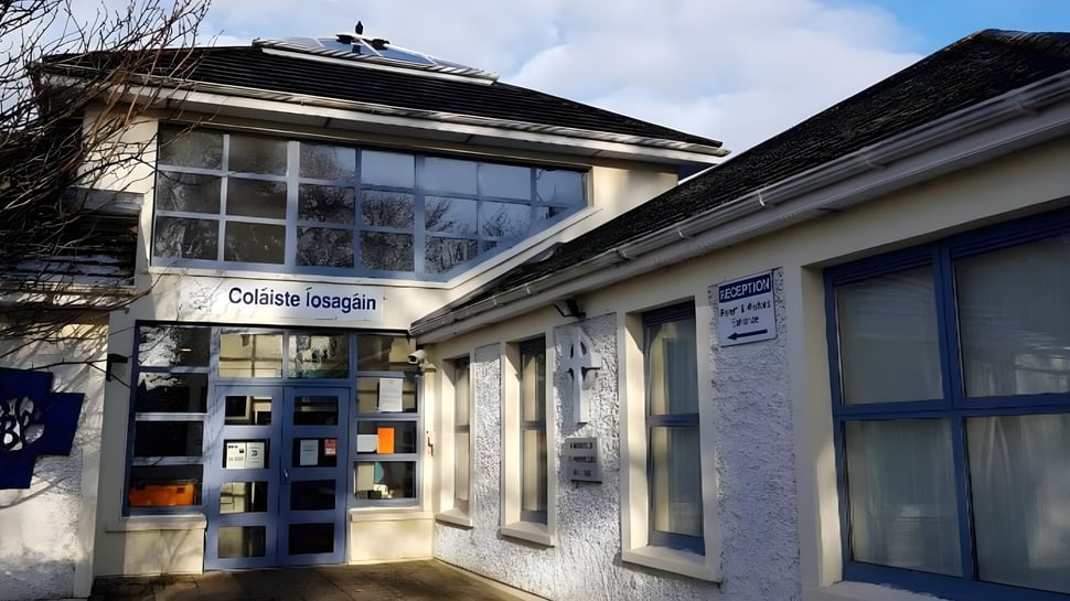 Das Gebäude von Coláiste Íosagáin ist aus Stein mit einem geneigten Dach und großen Fenstern unter bewölktem Himmel umgeben von kahlen Bäumen.