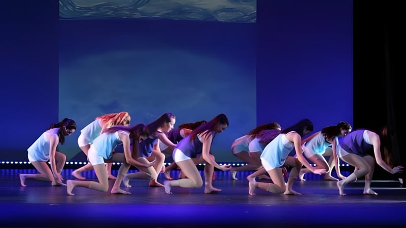 Eine Gruppe von Tänzerinnen und Tänzern führt auf der Bühne der Coláiste Mhuire Ballygar mit bunten Kostümen eine Tanzaufführung vor blauem Hintergrund auf.