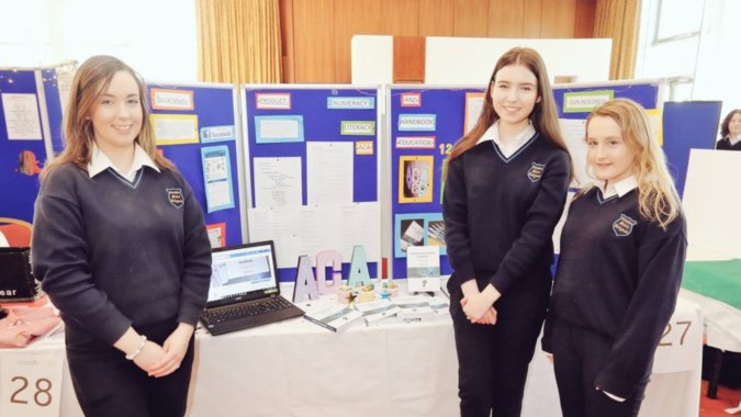 Drei Schülerinnen in Schuluniform stehen vor einem Informationsstand auf dem Campus von Coláiste Mhuire Ballymote.