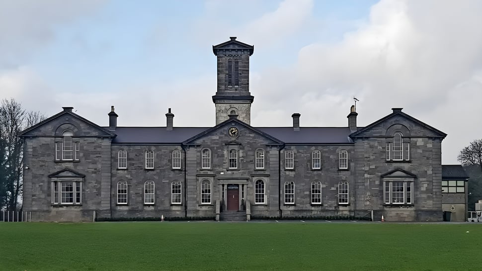 Das Gebäude mit einem hohen zentralen Turm der Coláiste Mhuire Mullingar steht auf einem grasbewachsenen Feld unter bewölktem Himmel.