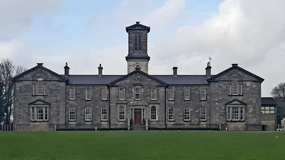 Das Gebäude mit einem hohen zentralen Turm der Coláiste Mhuire Mullingar steht auf einem grasbewachsenen Feld unter bewölktem Himmel.