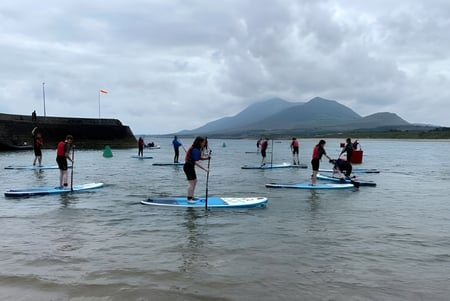 Schüler der Colaiste Mhuire (Tourmakeady) stehen auf Paddleboards in einem Gewässer mit Bergen im Hintergrund.