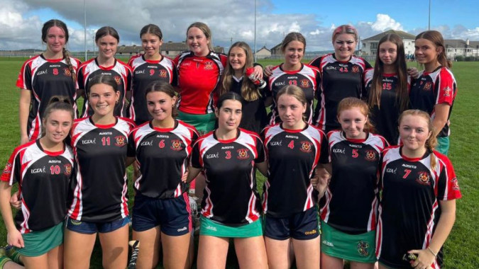 Eine Gruppe junger Sportlerinnen posiert auf dem Sportplatz der Colaiste Muire (Cobh) mit Schulgebäuden im Hintergrund.