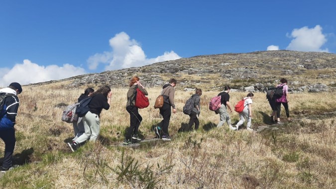 Eine Gruppe von Menschen geht auf einem grasbewachsenen Hügel unter blauem Himmel mit weißen Wolken auf dem Gelände von Coláiste Muire Crosshaven.