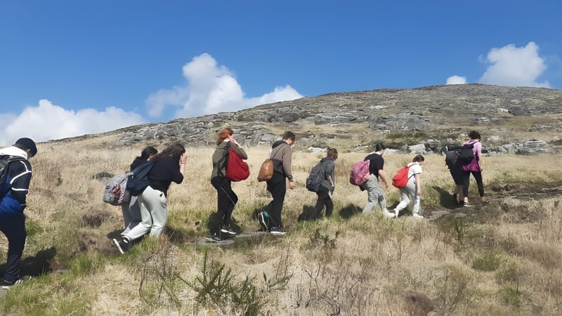 Eine Gruppe von Schülern der Coláiste Muire Crosshaven wandert auf einem grasbewachsenen Pfad in einer bergigen Landschaft unter blauem Himmel mit Wolken.
