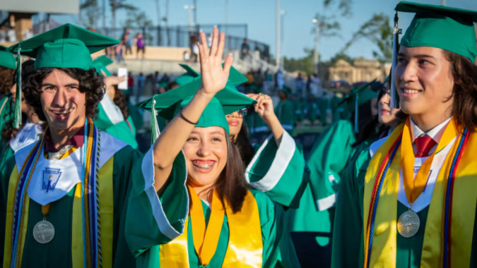 Eine Gruppe von Absolventen der Coláiste Naomh Cormac in grünen und gelben Talaren steht im Freien vor Gebäuden und Bäumen.