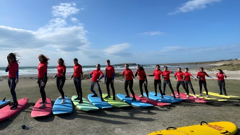 Schüler der Coláiste Pobail Acla stehen in roten Neoprenanzügen am Strand neben bunten Surfbrettern mit Küstenlandschaft im Hintergrund.