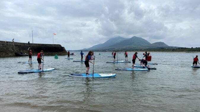 Eine Gruppe von Schülerinnen und Schülern der Colaiste Mhuire (Tourmakeady) steht auf Paddleboards auf einem Bergsee unter bewölktem Himmel.