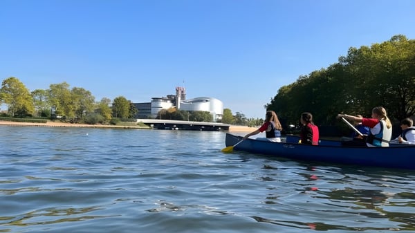 Eine Gruppe Schülerinnen und Schüler des Collège Episcopal Saint Étienne fährt mit einem blauen Boot auf einem Gewässer vor dem Schulgebäude.