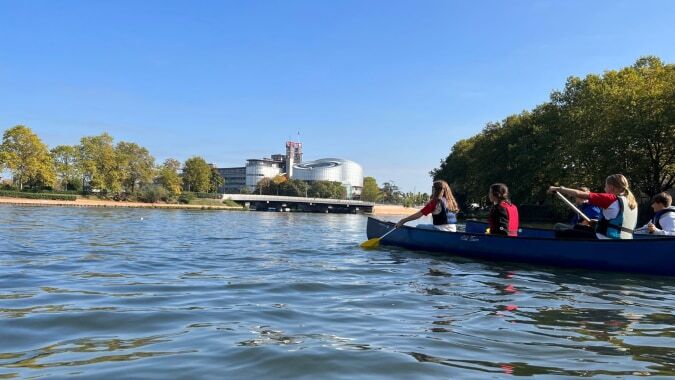 Schüler des Collège Episcopal Saint Étienne paddeln in einem blauen Kanu auf dem Wasser vor einem großen weißen Gebäude.
