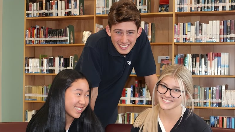 Drei junge Erwachsene stehen gemeinsam in der Bibliothek auf dem Campus des Collège Notre-Dame zwischen Bücherregalen.