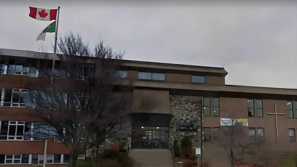 Das mehrstöckige Gebäude des Collège Notre-Dame mit kanadischer und palästinensischer Fahne auf dem Dach vor bewölktem Himmel.