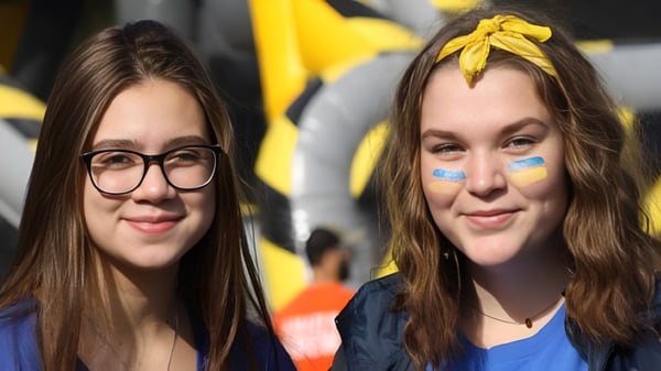 Zwei Schülerinnen des Collège Notre-Dame mit braunen Haaren und Gesichtsbemalung posieren vor einem bunten Hintergrund.