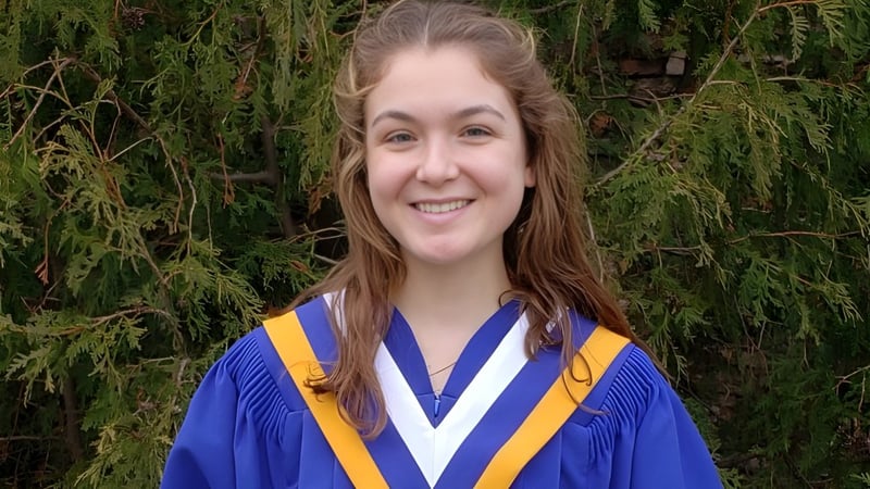 Eine junge Frau in blauen und gelben Abschlussgewand steht vor grünem Hintergrund auf dem Campus des Collège Sacré-Coeur.