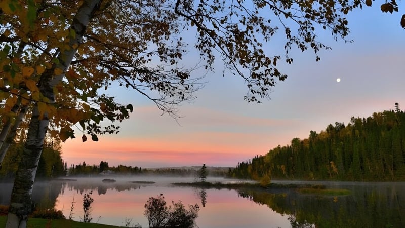 Ein See mit herbstlich gefärbten Bäumen und einem Halbmond am Himmel nahe dem College Saint-Bernard.