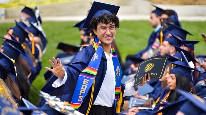 Ein Absolvent in blauem Talar und Barett steht mit weiteren Absolventen auf dem Campus des College of the Canyons im Freien.