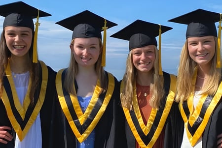 Vier Schülerinnen in Abschlusskleidung stehen vor blauem Himmel auf dem Campus des Collingwood Collegiate Institute.