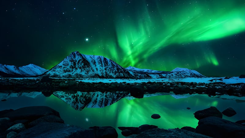 Das Bild zeigt die Aurora Borealis über einem stillen See mit reflektierten schneebedeckten Bergen bei Nacht.