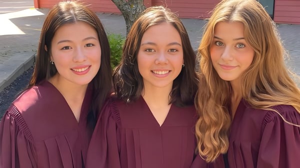 Drei Absolventinnen in Talaren posieren vor einem roten Holzgebäude auf dem Campus der Colonel Gray High School.