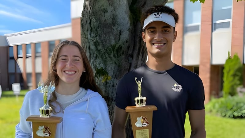 Zwei Personen stehen vor einem Baum auf dem Campus der Colonel Gray High School und halten Trophäen in der Hand.