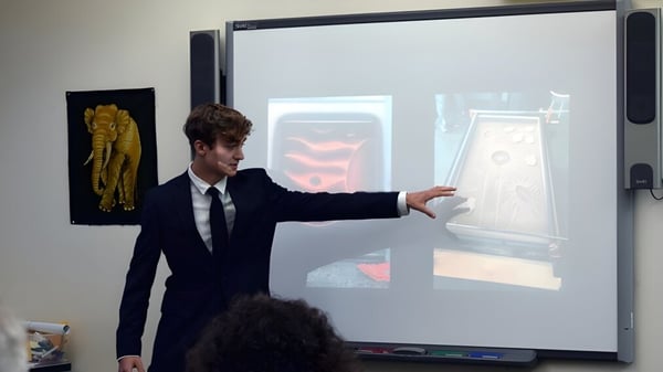 Ein Mann im Anzug steht vor einer großen Leinwand und zeigt auf eine Projektion auf dem Campus der Colorado Rocky Mountain School.