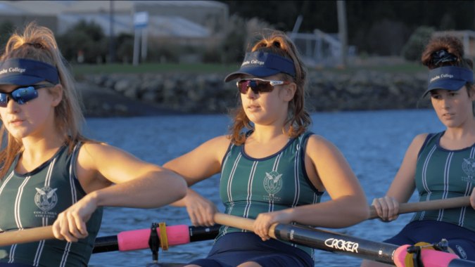 Drei Schülerinnen des Columba College stehen in Sportbekleidung nahe einem Gewässer mit Bäumen und Bergen im Hintergrund.