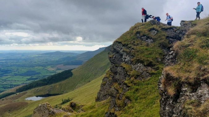Eine Gruppe von Menschen steht auf einer Felsklippe mit Blick auf ein grünes Tal und Berge auf dem Gelände des Comeragh College.