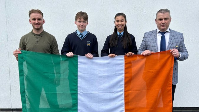Zwei Männer und zwei Jugendliche halten vor neutralem Hintergrund die Flagge von Irland am Comeragh College.