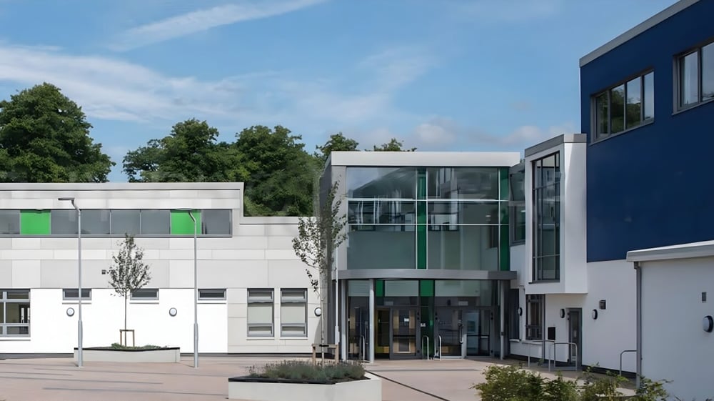 Das moderne mehrstöckige Gebäude des Comeragh College mit Glaseingang und grünen sowie blauen Akzenten von Bäumen umgeben.