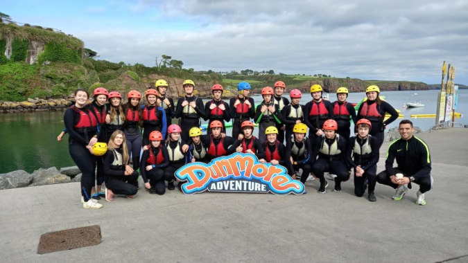 Eine Gruppe von Schülerinnen und Schülern des Comeragh College in roten und schwarzen Neoprenanzügen steht vor einer Küstenlandschaft mit Klippen und Wasser.