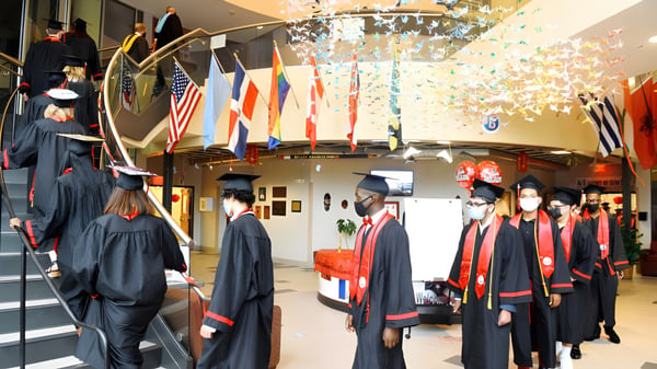 Absolventinnen und Absolventen der Connecticut IB Academy in schwarzen Talaren gehen durch einen Flur mit internationalen Fahnen während der Abschlussfeier.