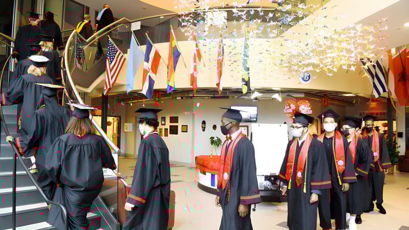 Absolventinnen und Absolventen der Connecticut IB Academy in schwarzen Talaren gehen durch einen Flur mit internationalen Fahnen während der Abschlussfeier.