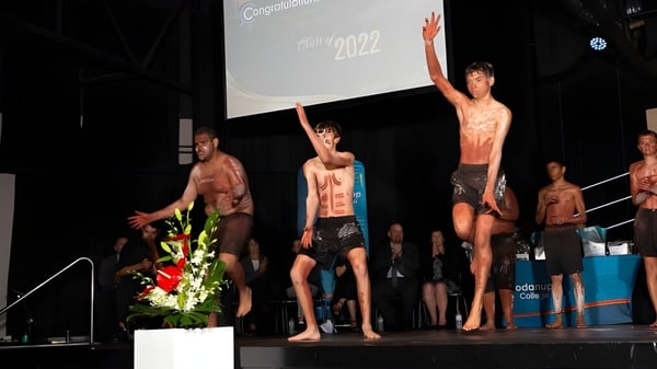 Eine Gruppe von jungen Männern steht ohne Hemd auf der Bühne auf dem Campus des Coodanup College mit einer großen Leinwand im Hintergrund, die das Jahr 2022 zeigt.