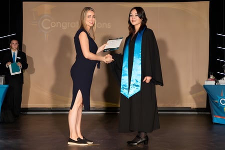 Zwei Frauen im formellen Outfit geben sich auf der Bühne die Hand vor einem Glückwunsch-Banner auf dem Campus der Coodanup College.