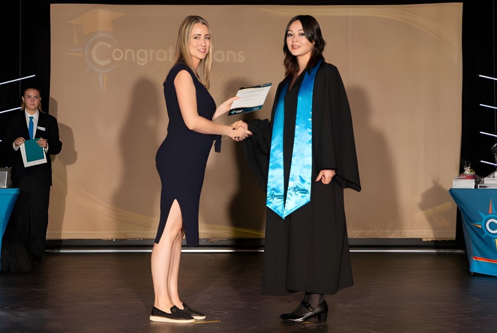 Zwei Frauen im formellen Outfit geben sich auf der Bühne die Hand vor einem Glückwunsch-Banner auf dem Campus der Coodanup College.