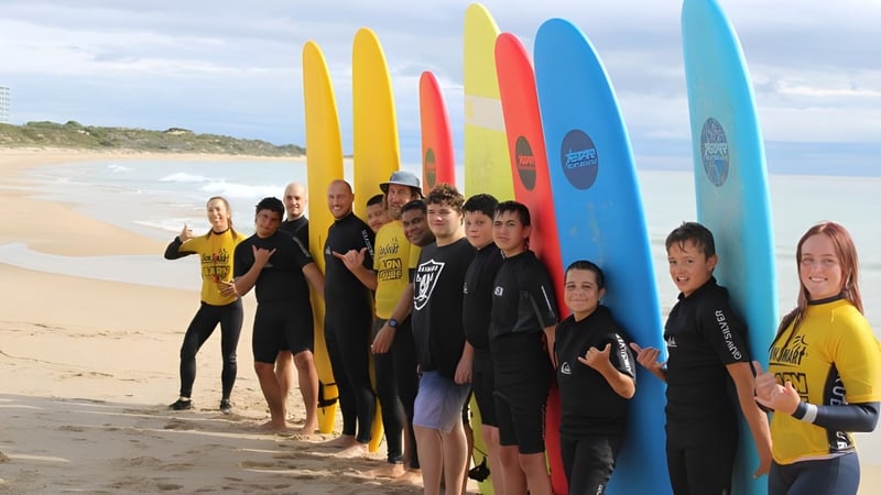 Schüler des Coodanup College stehen in Neoprenanzügen am Strand mit bunten Surfbrettern vor einer Küstenlandschaft.
