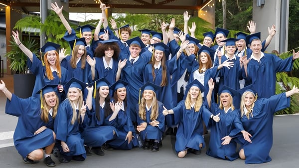 Eine große Gruppe von Absolventen feiert in blauen Talaren und Hüten auf dem Campus der Coolum State High School.