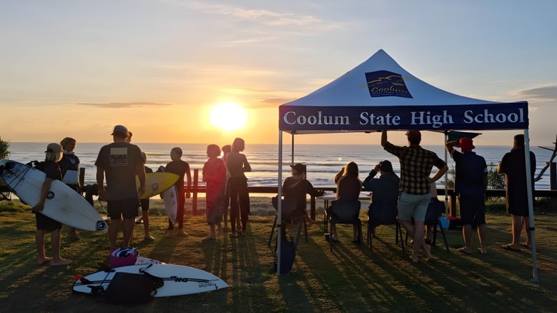 Eine Gruppe Schüler steht vor dem Coolum State High School Banner am Strand bei Sonnenuntergang.