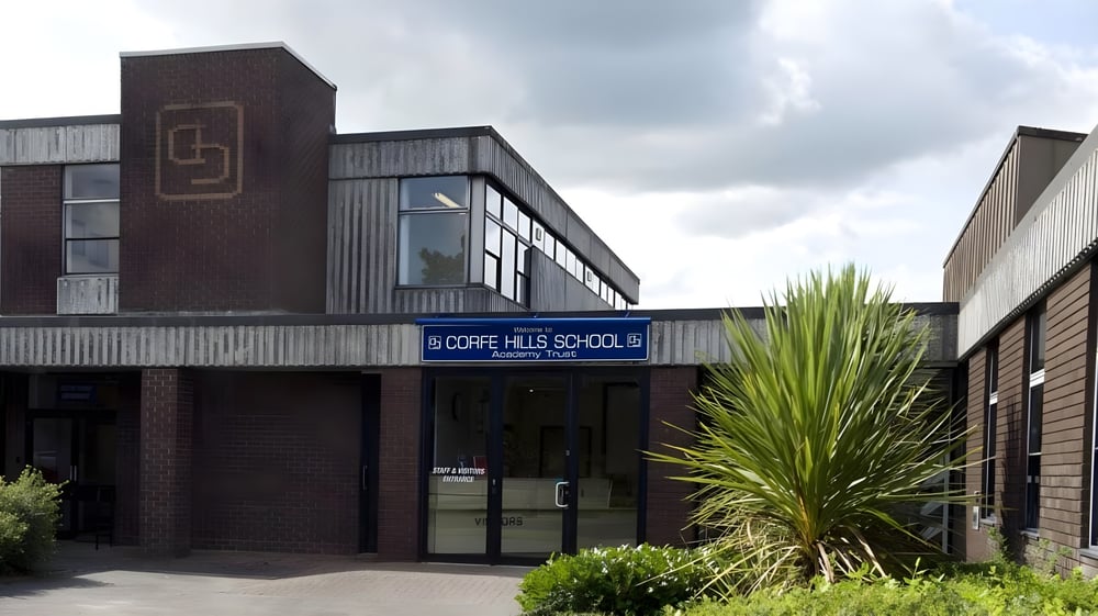 Das moderne Gebäude der Corfe Hills School mit großen Fenstern und einer Palme im Vordergrund unter bewölktem Himmel.