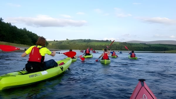 Schüler der Cork Educate Together Secondary School paddeln mit bunten Kajaks auf einem ruhigen See inmitten grüner Wälder.