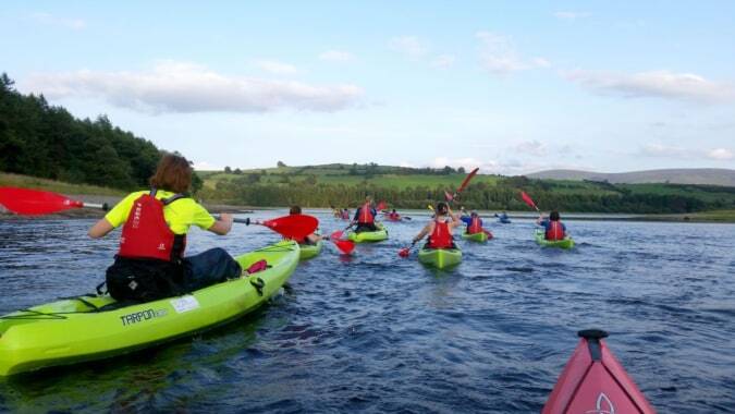 Schüler der Cork Educate Together Secondary School paddeln mit bunten Kajaks auf einem ruhigen See vor grünen Wäldern.