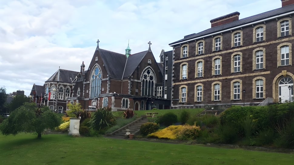 Das historische Gebäude der Cork Educate Together Secondary School mit einem gepflegten Garten im Vordergrund unter bewölktem Himmel.