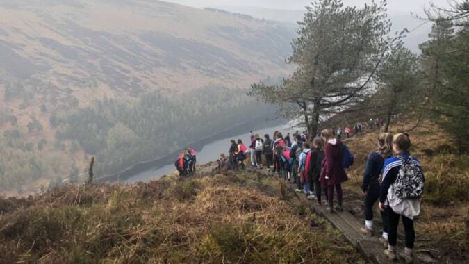 Eine Gruppe von Schülerinnen und Schülern der Cornafulla National School wandert auf einem Weg entlang einer bergigen Landschaft mit Fluss im Hintergrund.
