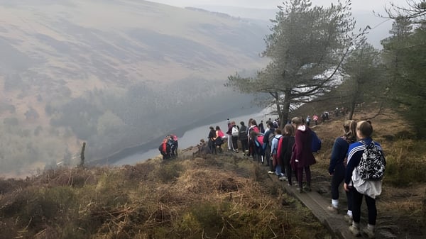 Eine Gruppe von Schülerinnen und Schülern der Cornafulla National School wandert auf einem Pfad durch eine bergige Waldlandschaft mit Fluss im Hintergrund.