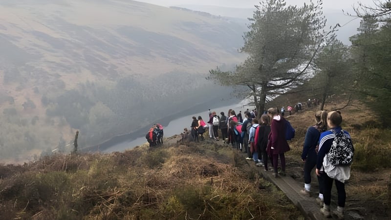 Eine Gruppe von Schülerinnen und Schülern der Cornafulla National School wandert auf einem Pfad durch eine bergige Waldlandschaft mit Fluss im Hintergrund.