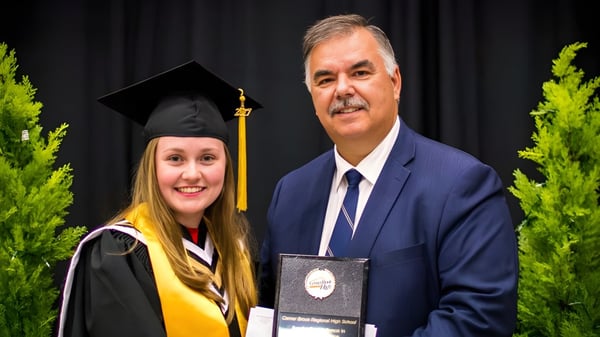 Eine Absolventin steht neben einem Mann im Anzug vor einer grünen Kulisse auf dem Gelände der Corner Brook Regional High School.