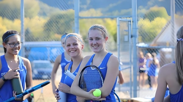 Drei Schülerinnen in Sportkleidung mit Tennisschlaegern stehen vor dem Tennisplatz der Cotter Schools vor einer Bergkulisse.